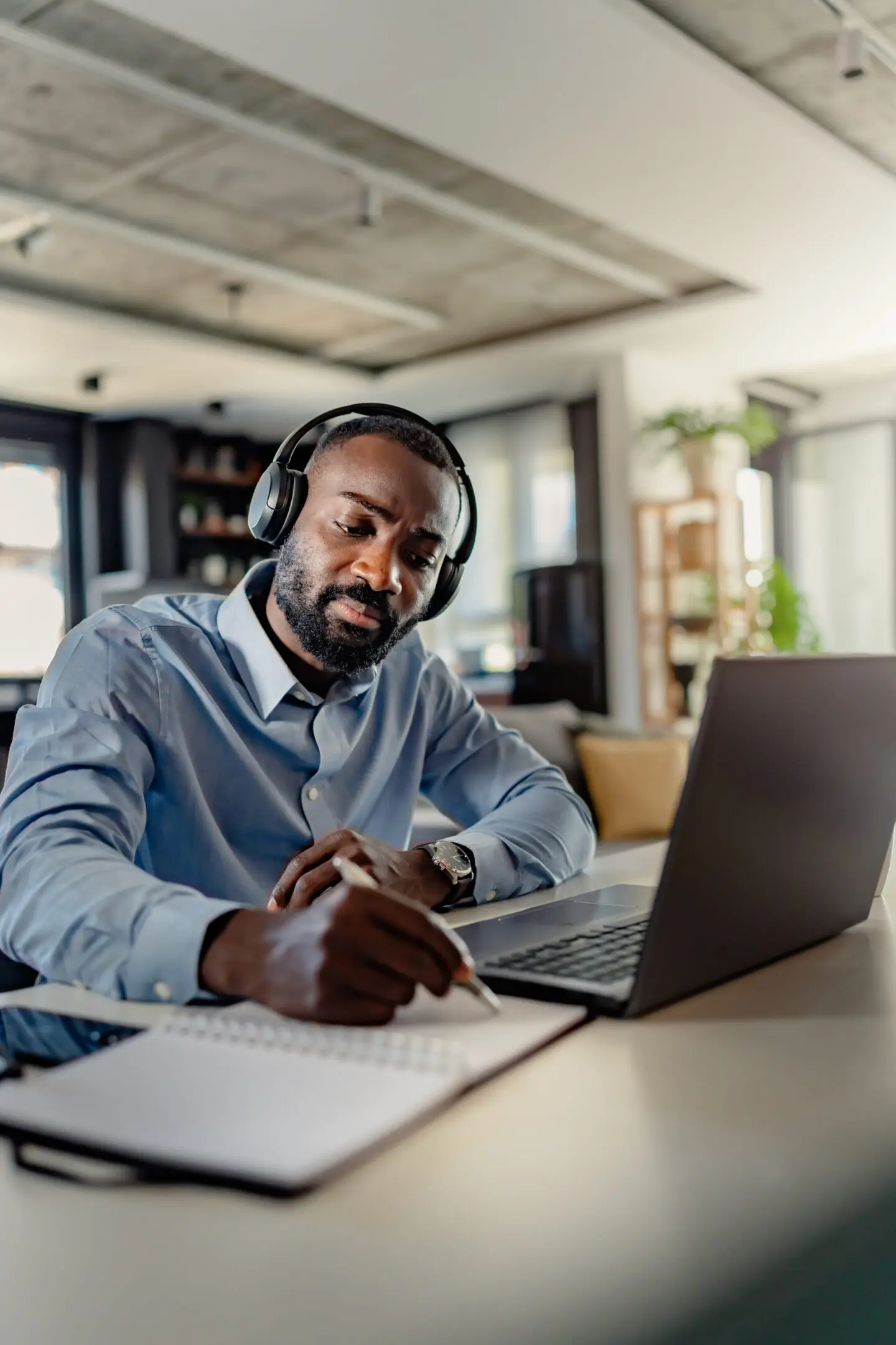Man with headphones writing notes while using a laptop.